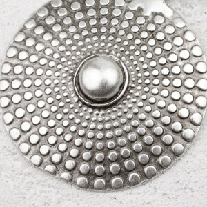 Puede incluir: Un colgante de metal plateado con un diseño circular. El colgante tiene un centro elevado con un patrón circular de pequeños puntos que irradian hacia afuera.