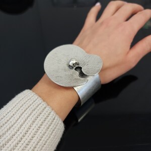 May include: A silver cuff bracelet with a textured, circular design and a small silver ball in the center.