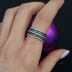 May include: A silver ring with a braided design. The ring is worn on a finger and the background is a purple orb.