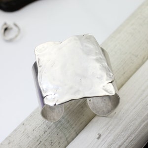 Op de afbeelding: Een zilveren armband met een getextureerd, gehamerd, vierkant ontwerp. De armband is aan &eacute;&eacute;n kant open en heeft een gladde, afgeronde rand.