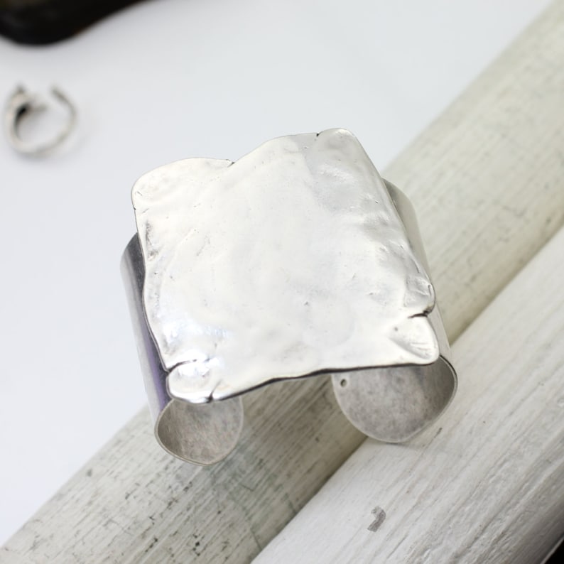 Op de afbeelding: Een zilveren armband met een getextureerd, gehamerd, vierkant ontwerp. De armband is aan &eacute;&eacute;n kant open en heeft een gladde, afgeronde rand.