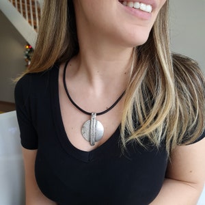 Op de afbeelding: Een zilveren ketting met een ronde, getextureerde hanger en een verticaal balkontwerp. De ketting is aan een zwart koord.
