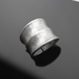 Könnte beinhalten: Ein silberfarbener Ring mit einem breiten, strukturierten Band. Der Ring hat eine leicht gebogene Form und eine verstellbare Öffnung. Das Metall hat eine matte Oberfläche, die ihm ein Vintage- oder handgefertigtes Aussehen verleiht. Der Ring ist als Modeaccessoire konzipiert.