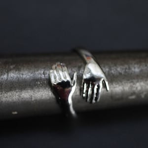 Pode incluir: Um anel de prata com duas mãos entrelaçadas, uma mão estendida e a outra a segurando.