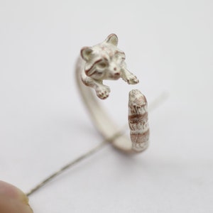 May include: A silver ring with a raccoon design. The raccoon is curled up with its tail wrapped around the ring band. The ring is a delicate piece of jewelry.