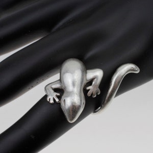 May include: A silver ring featuring a detailed lizard design. The lizard is curled around the ring band, with its tail extending to the side.