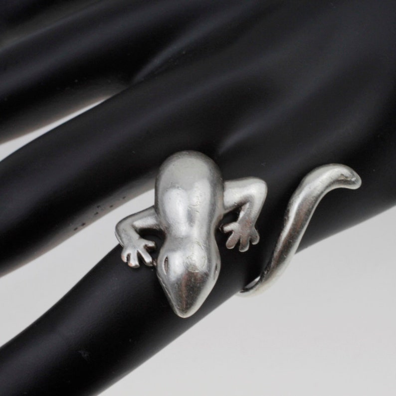 May include: A silver ring featuring a detailed lizard design. The lizard is curled around the ring band, with its tail extending to the side.