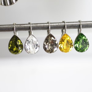 Puede incluir: Una fila de cinco pendientes en forma de lágrima, en varios colores: verde oliva, transparente, gris ahumado, amarillo y verde esmeralda. Cada pendiente tiene un engaste y un gancho de metal plateado. Los pendientes se muestran sobre una barra plateada.