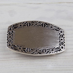 May include: A silver belt buckle with a decorative, ornate design featuring a repeating pattern of flowers and stars. The buckle has a blank, rectangular space in the centre.
