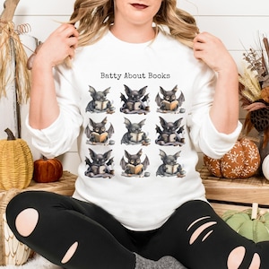 May include: White long-sleeved sweatshirt with the text "Batty About Books" above a pattern of watercolor bats reading books. The bats are gray with black wings and are holding open books.