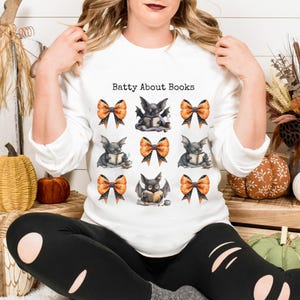 May include: White long-sleeved sweatshirt with the text "Batty About Books". The design features illustrations of bats reading books, interspersed with orange and black polka-dot bows. The overall theme is Halloween and reading.