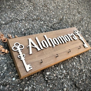 May include: Wooden key holder with the text "Alohomora" in a decorative font. The holder has four hooks and two key silhouettes.