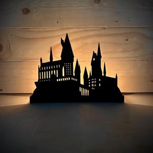 Puede incluir: Una silueta negra del castillo de Hogwarts, una escuela ficticia de brujería y magia, está iluminada desde atrás. La silueta está hecha de madera y es una pieza decorativa.
