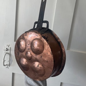 May include: Antique copper pan with a black iron handle. The pan has five round indentations in the bottom, likely for cooking eggs.