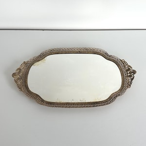 May include: An antique oval vanity mirror with a decorative gold-toned filigree frame. The mirror has a slightly aged appearance, adding to its vintage charm. A classic piece for a dressing table or vanity.