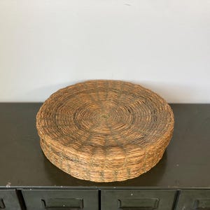 May include: A round woven straw cushion with a natural brown color. The cushion is made of tightly woven straw and has a circular shape.