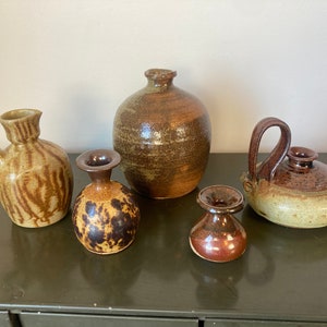 May include: A collection of handcrafted ceramic vases and vessels in various shapes and sizes. The pottery features earthy tones of brown, tan, and speckled patterns. One vase has a handle, and another has a unique, rounded shape.