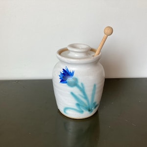 May include: A white ceramic jar with a lid and a wooden honey dipper. The jar is decorated with a blue floral design. The honey dipper is made of light-colored wood.