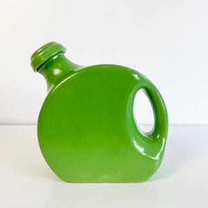 May include: A bright lime green ceramic decanter with a circular, flattened body, a side handle, and a matching stopper. The vessel features a smooth, glossy finish and a distinct, rounded silhouette that sits flat on its base.