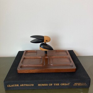 May include: A wooden tray with four compartments and a carved wooden bird figure in the centre. The tray is brown and the bird is black and brown. The tray is sitting on top of a black book with gold lettering that reads "Claude Arthaud Homes of the Great Weidenfeld and Nicolson."