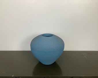 May include: A blue ceramic vase with a textured surface. The vase has a wide opening and a rounded shape.