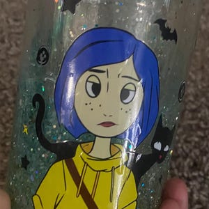 Puede incluir: Vaso lleno de purpurina con una ilustraci&oacute;n de dibujos animados de una ni&ntilde;a con pelo azul y una chaqueta amarilla, junto con una silueta de gato negro. El vaso tiene una tapa verde azulado y est&aacute; decorado con estrellas, murci&eacute;lagos y lunas crecientes.