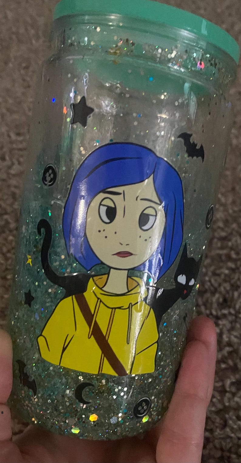 Puede incluir: Vaso lleno de purpurina con una ilustraci&oacute;n de dibujos animados de una ni&ntilde;a con pelo azul y una chaqueta amarilla, junto con una silueta de gato negro. El vaso tiene una tapa verde azulado y est&aacute; decorado con estrellas, murci&eacute;lagos y lunas crecientes.