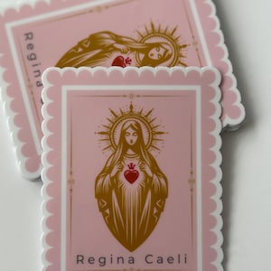 Può includere: Adesivo rettangolare rosa con bordo smerlato. Presenta un'illustrazione dorata di una figura religiosa con un cuore e un'aureola, e la scritta "Regina Caeli". Un altro adesivo è parzialmente visibile sullo sfondo.