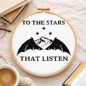 以下が含まれることがあります： 「TO THE STARS THAT LISTEN」の文字が黒で刺繍されたクロスステッチの刺繍枠。デザインには山、星、コウモリのような翼が含まれています。枠は、金色の留め金が付いた木製のフレームにセットされています。