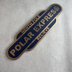 May include: Wooden sign with a navy blue background and gold lettering that reads "POLAR EXPRESS" and "NORTH POLE BELIEVE". The sign has a gold border and is in a layered design.