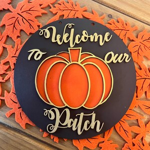 May include: A round wooden sign with a brown background and gold lettering. The sign says "Welcome to our Patch" and features an orange pumpkin in the centre.