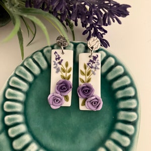 Puede incluir: Un par de pendientes rectangulares blancos colgantes con flores moradas y hojas verdes. Los pendientes tienen ganchos plateados.