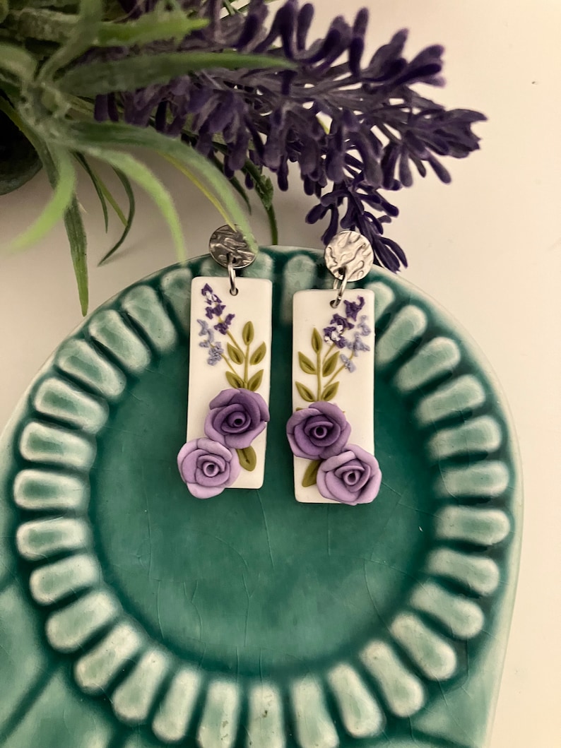 Puede incluir: Un par de pendientes rectangulares blancos colgantes con flores moradas y hojas verdes. Los pendientes tienen ganchos plateados.