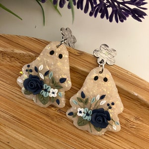 May include: A pair of ghost-shaped earrings with a floral design. The earrings are made of white polymer clay with a glittery finish and feature blue and white flowers.