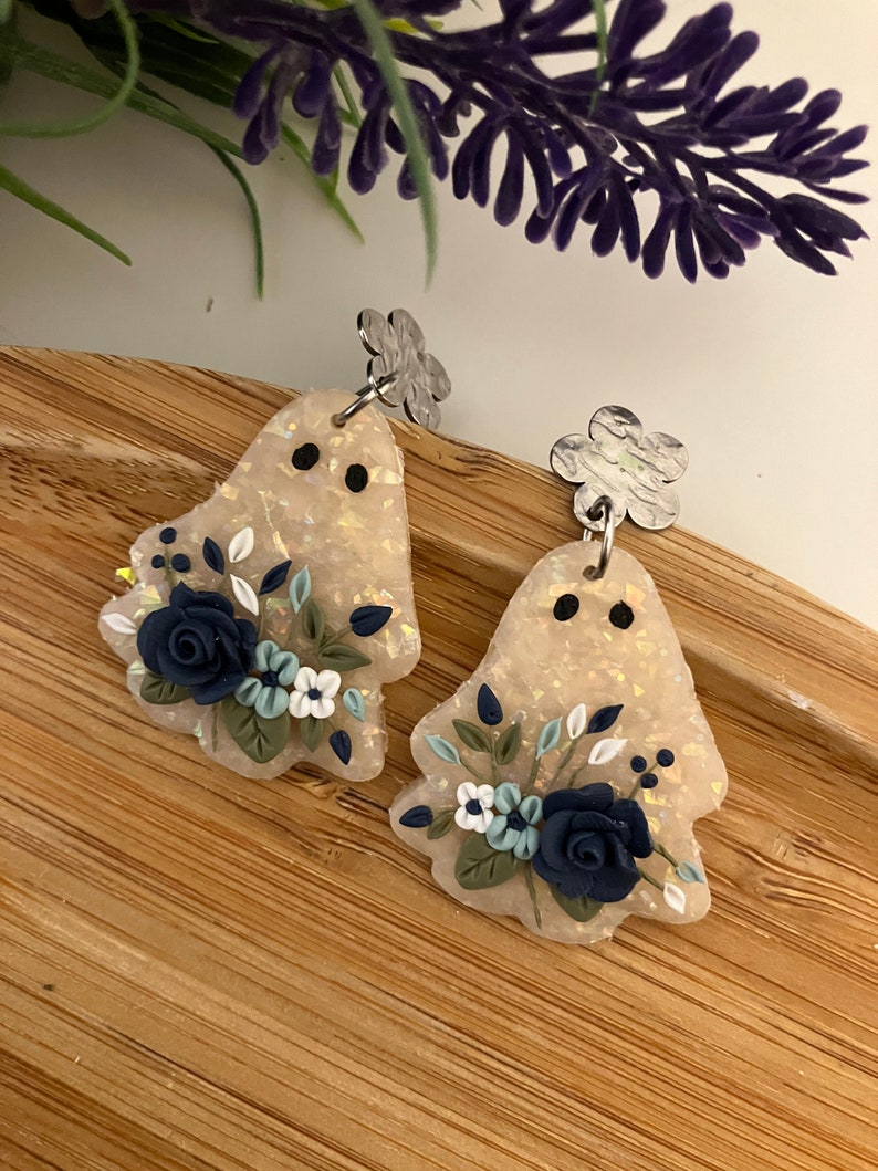 May include: A pair of ghost-shaped earrings with a floral design. The earrings are made of white polymer clay with a glittery finish and feature blue and white flowers.
