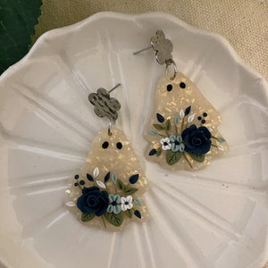 May include: A pair of white and gold iridescent dangle earrings with a floral design. The flowers are blue, white, and green. The earrings have a silver flower charm at the top.