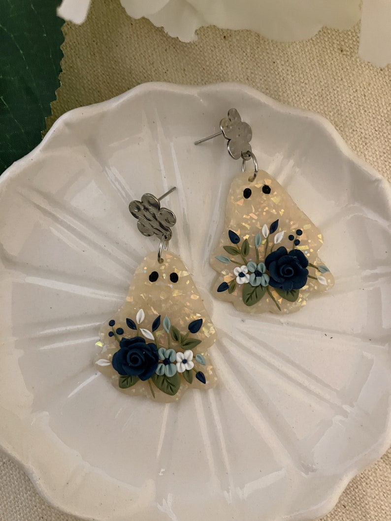 May include: A pair of white and gold iridescent dangle earrings with a floral design. The flowers are blue, white, and green. The earrings have a silver flower charm at the top.