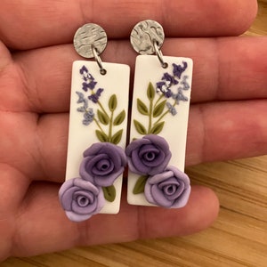 Puede incluir: Un par de pendientes rectangulares blancos colgantes con flores moradas y hojas verdes. Los pendientes tienen ganchos de metal plateado.