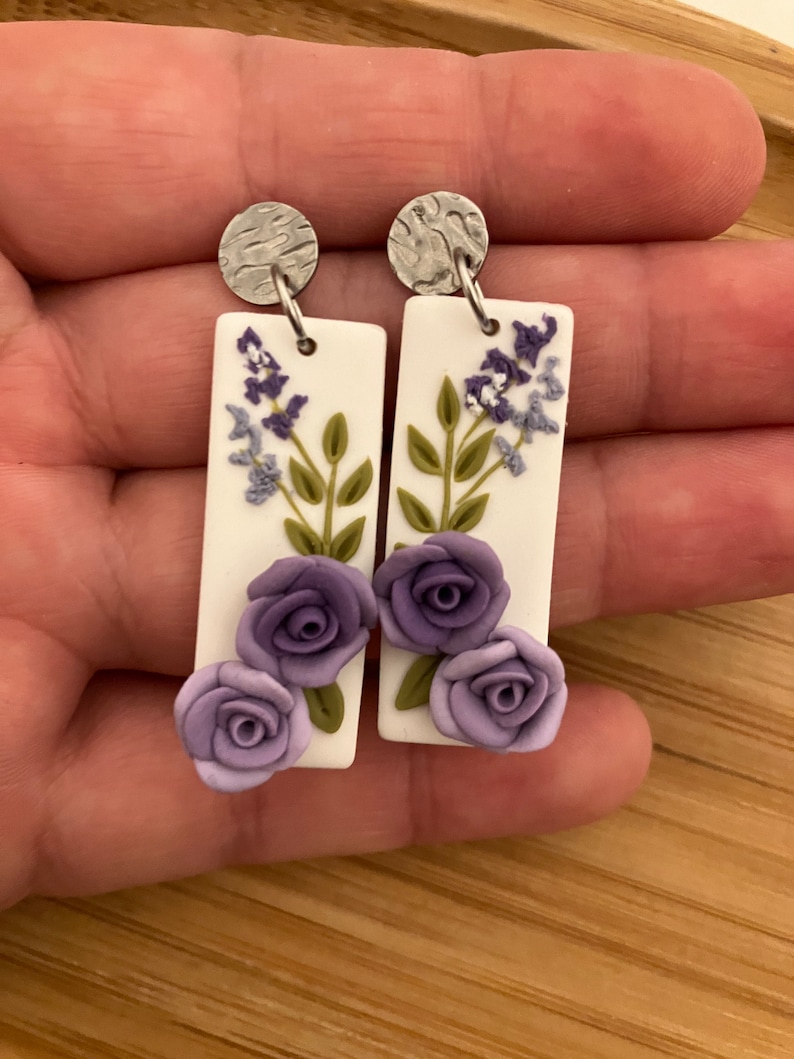 Puede incluir: Un par de pendientes rectangulares blancos colgantes con flores moradas y hojas verdes. Los pendientes tienen ganchos de metal plateado.