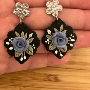 May include: A pair of black and silver earrings with a blue flower design. The earrings are made of polymer clay and have a silver flower charm at the top.