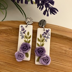 Puede incluir: Un par de pendientes rectangulares blancos colgantes con dise&ntilde;os florales morados y verdes. Los pendientes tienen ganchos de metal plateado.