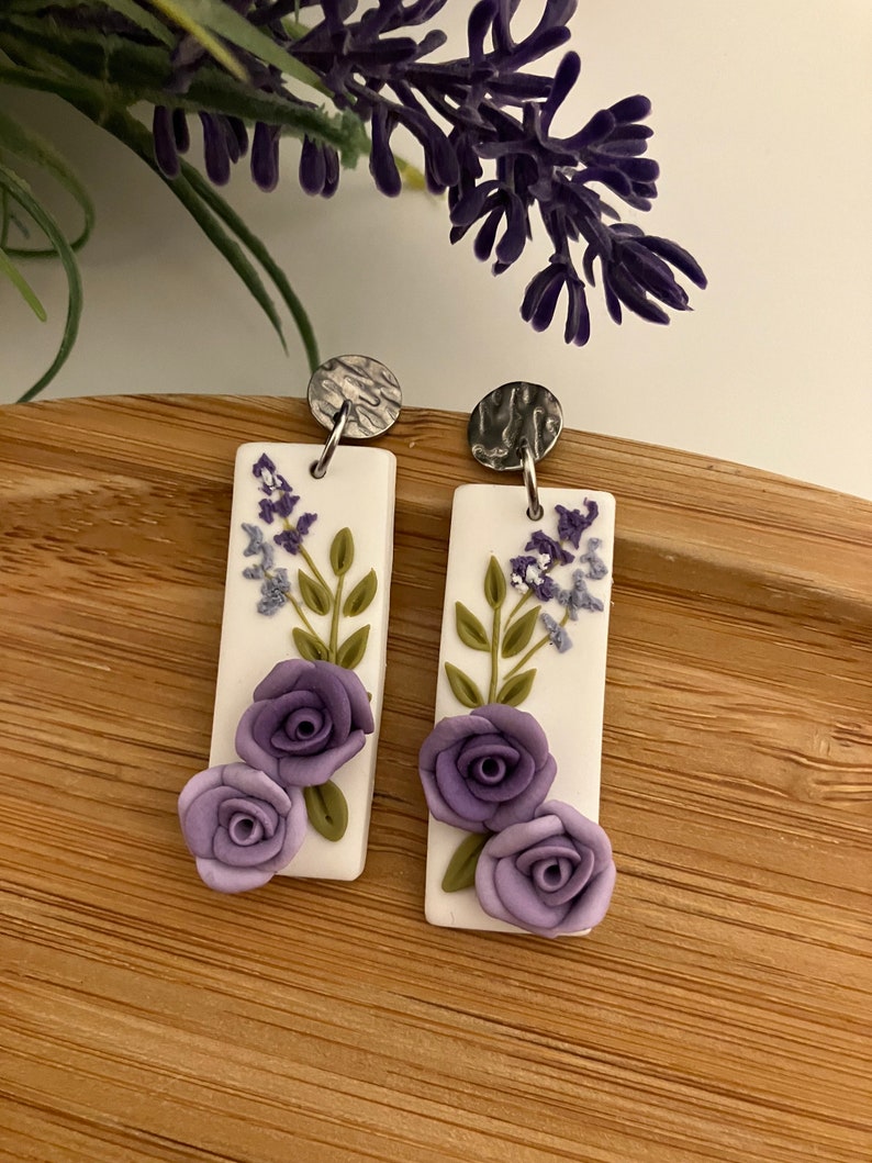 Puede incluir: Un par de pendientes rectangulares blancos colgantes con dise&ntilde;os florales morados y verdes. Los pendientes tienen ganchos de metal plateado.