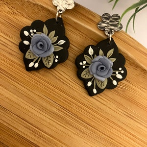 May include: A pair of black polymer clay earrings with a floral design. Each earring features a blue flower with green leaves and white accents. The earrings are attached to silver-toned metal hooks.
