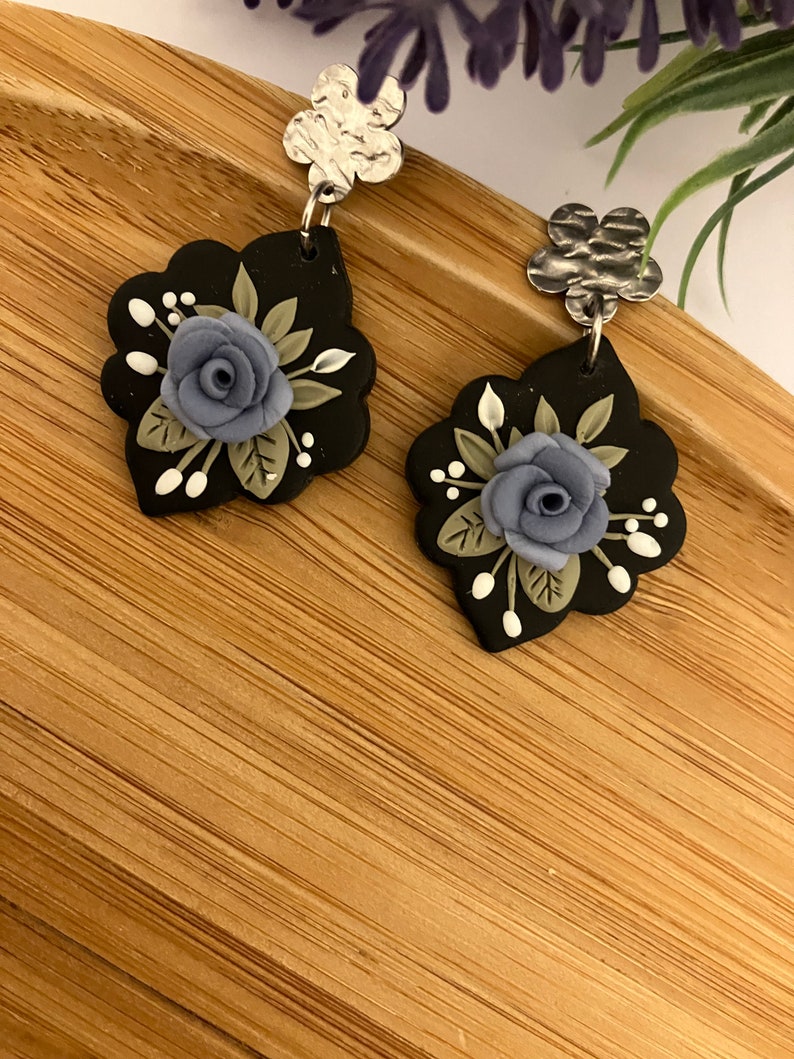 May include: A pair of black polymer clay earrings with a floral design. Each earring features a blue flower with green leaves and white accents. The earrings are attached to silver-toned metal hooks.