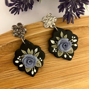 May include: A pair of black and silver earrings with a floral design. The earrings feature a blue flower with green leaves and white accents.