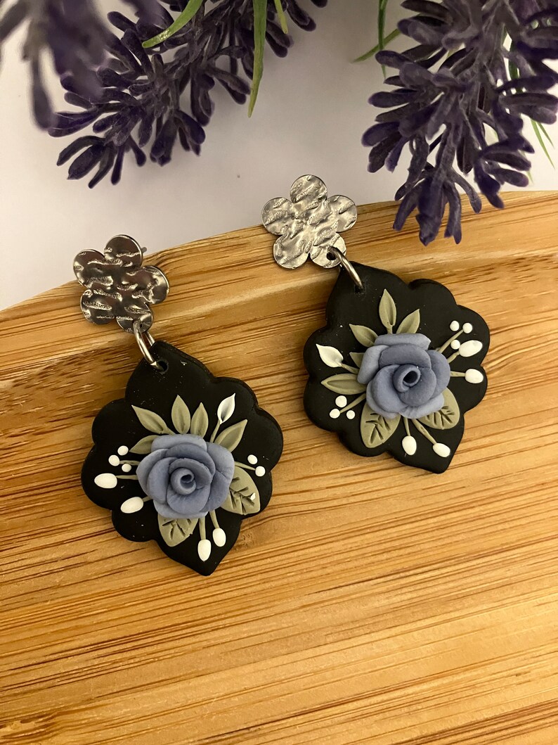 May include: A pair of black and silver earrings with a floral design. The earrings feature a blue flower with green leaves and white accents.