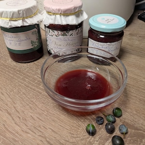 Könnte beinhalten: Drei Gläser hausgemachter Konfitüre mit handgeschriebenen Etiketten und dekorativen Deckeln. Eine kleine Glasschale enthält einen Teil der Konfitüre, und mehrere dunkle Beeren sind in der Nähe verstreut. Die Gläser sind mit Namen und Zahlen beschriftet.