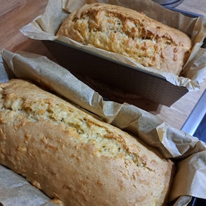 Könnte beinhalten: Zwei frisch gebackene Brote in braunen Brotformen, die mit Backpapier ausgelegt sind. Das Brot ist goldbraun und hat eine leicht raue Textur.