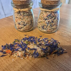 Könnte beinhalten: Zwei kleine Glasgefäße mit Korkdeckeln, gefüllt mit einer Mischung aus Salz, getrockneten Kräutern und blauen Blütenblättern. Die Gläser stehen auf einer Holzoberfläche, und vor ihnen liegt ein Haufen der Salzmischung.
