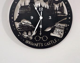 May include: A circular black wall clock featuring laser-cut silhouettes of Hogwarts Castle, a steam train, a broomstick, and round glasses. The text Harry Potter and HOGWARTS CASTLE is engraved. It includes white clock hands and numbers 3, 6, 9, and 12.